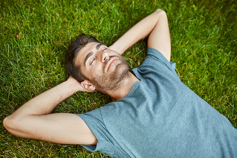 Portrait of young mature good-looking caucasian man in blue shirt peaceful lying on grass with yes closed. Guy sleeping in garden near country house in peaceful summer day.